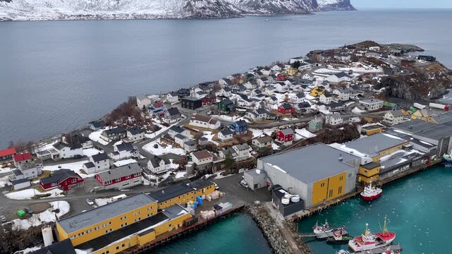 Aerial view of the coastal village of Husoy with colorful houses, fishing boats, and snow-capped mountains in the background in Husoy, Troms, Norway.