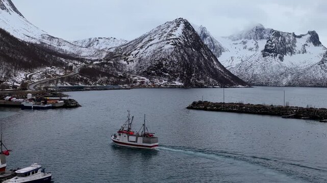 Aerial view of a fishing boat navigating the calm waters of the harbor surrounded by snow-capped mountains in Husoy, Troms, Norway.