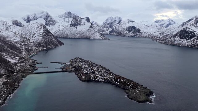 Aerial view of the village of Husoy situated on an island surrounded by snow-capped mountains and deep blue fjords in Husoy, Troms, Norway.