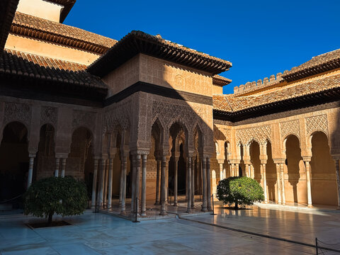 View of the intricate Moorish architecture of the Alhambra courtyard with ornate arches, marble columns, and manicured trees in Granada, Andalucia, Spain.