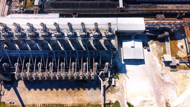Aerial view of heavy industrial machinery and processing pipes. Steel pipelines and processing silos stand on concrete ground in bright sunlight.