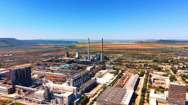 Panoramic aerial view of an expansive industrial complex. Factory buildings, pipelines, and smokestacks stretch toward distant hills under clear summer skies.