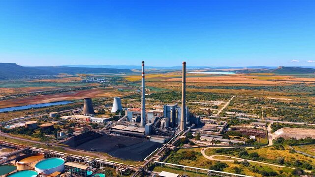 Wide aerial view of power plant with tall chimneys. Two tall smokestacks and cooling towers stand near water basins in an open landscape under blue sky.
