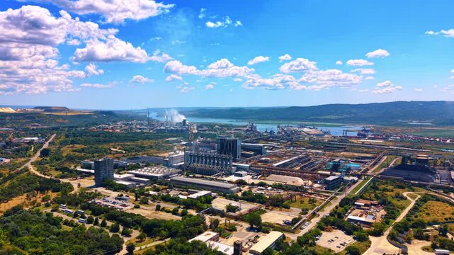 Sprawling industrial site surrounded by green trees and parking lots. Factory buildings and storage areas spread across a wide landscape under a cloudy blue sky.
