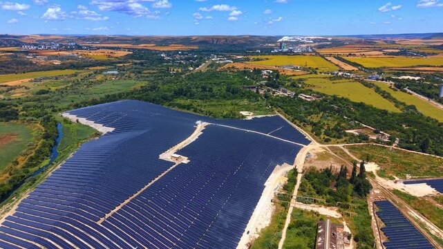Solar panel farm on countryside slope aerial view. A large array of dark panels sits on rolling hills near green vegetation.