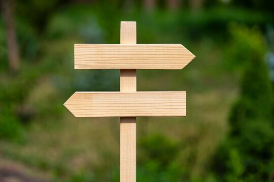 Blank wooden directional signpost with arrows pointing opposite ways, closeup. Empty wooden sign post with two arrows pointing left and right on blurry green nature background in summer garden
