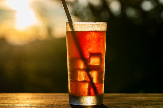 Iced tea drink in glass at golden hour sunset, closeup. Refreshing glass of iced tea or cold brew coffee with ice cubes and straw on wooden table at sunset bokeh background