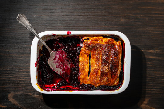 Homemade blueberry cobbler pie in baking dish top view, closeup. Half eaten traditional berry pie or cobbler in white ceramic baking pan with vintage serving spatula on dark wooden background