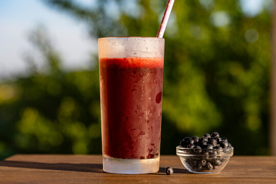 Fresh blueberry smoothie in glass, outdoors, closeup. Chilled berry smoothie or juice in frosted glass with paper straw next to small bowl of fresh blueberries on table over blurry green background