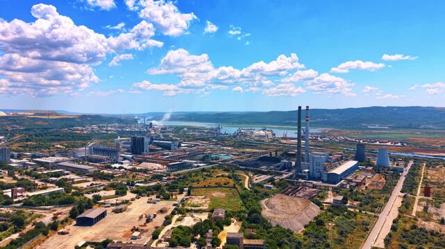 Panoramic aerial view of power station and industrial zone. Tall smokestacks and factory structures stand in a wide industrial valley near water under a bright sky.