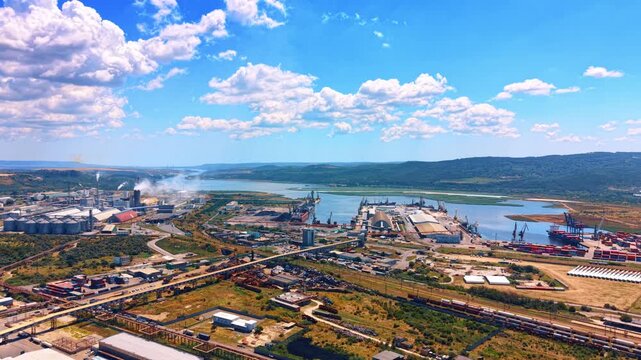 High angle aerial shot of seaport terminal and industrial district. Port docks, cargo shipping areas, and factory buildings extend toward a river and hills under a cloudy sky.