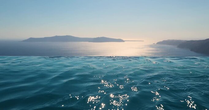 Turquoise blue water ripples in an infinity pool overlooking the caldera of Santorini, Greece as the sunset begins in the background. The wind is blowing. 