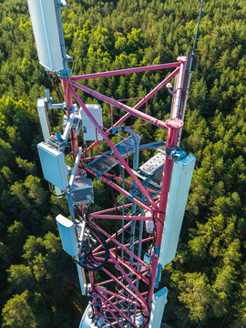 Aerial view of a telecommunications tower with red metal framework and multiple antennas rising above a dense green forest in Riga, Latvia.