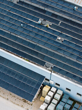 Aerial view of a factory roof covered with rows of solar panels, with stacks of concrete paving stones and construction materials stored in the yard below in Riga, Latvia.