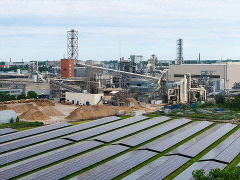 Aerial view of a manufacturing plant for wood-based materials featuring industrial structures, conveyor belts, piles of wood chips, and a large solar panel array in Riga, Latvia.
