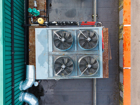 Aerial view of an industrial HVAC cooling unit with four large fans mounted on a flat rooftop, featuring metal piping and surrounding corrugated walls in Riga, Latvia.