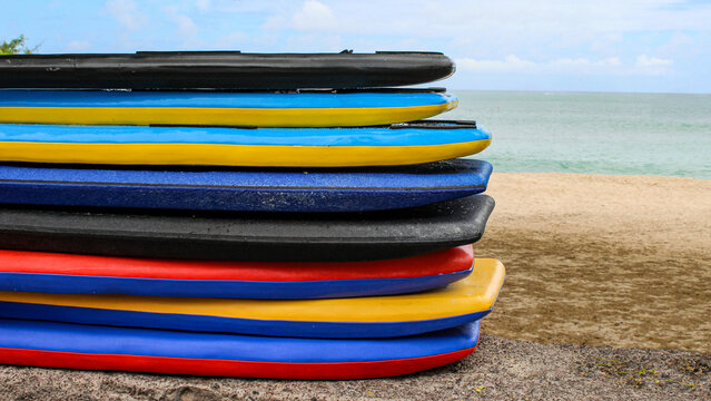 Stack of colorful stand-up paddleboards on a tropical sandy beach with boats turquoise ocean background.