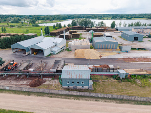 Aerial view of a timber processing facility featuring industrial buildings, stacks of logs, and wood chip piles near a lake in Brankas, Jelgava Municipality, Latvia.