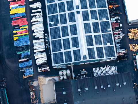 Aerial view of a polymer pipe manufacturing facility with a large warehouse roof covered in solar panels, rows of colorful plastic pipes, and industrial storage areas in Jelgava, Latvia.