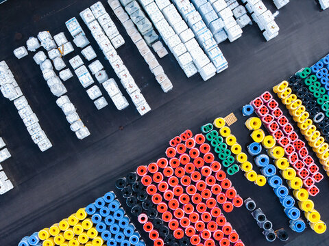 Aerial view of an industrial storage yard featuring organized stacks of white concrete blocks and rows of colorful plastic pipes in red, yellow, blue, green, and black, Jelgava, Latvia.