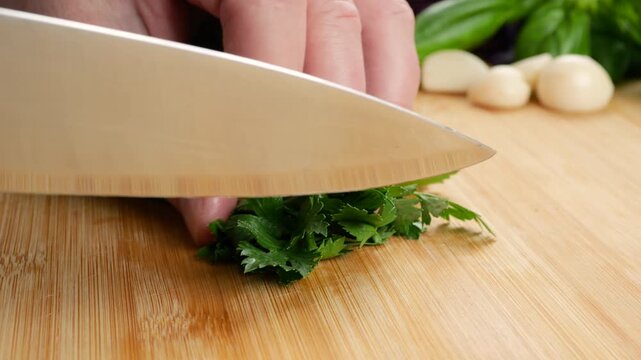 Chef finely chops fresh green parsley on a wooden cutting board with a sharp knife. Peeled garlic and basil rest in the blurred background, capturing organic ingredient prep in a kitchen.