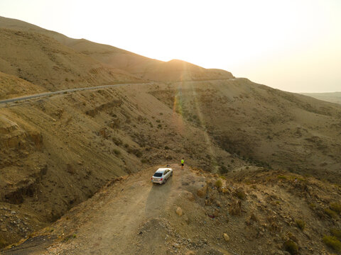 Aerial view of a car parked on a dirt ridge with a person standing nearby, overlooking the rugged desert landscape of the Maeen Sub-District in Machaerus, Madaba Governorate, Jordan.