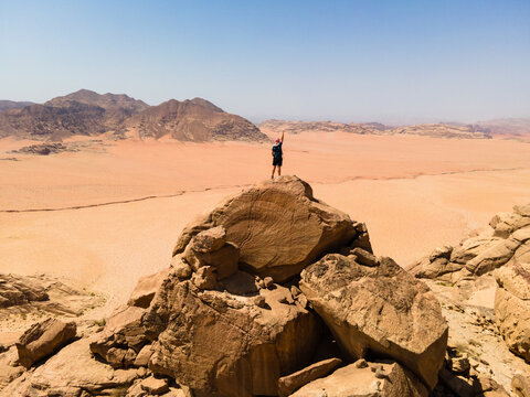 Aerial view of a hiker standing on a rock formation with arm raised in the vast desert landscape of Jabal Al-Hash in Wadi Rum, Aqaba, Aqaba Governorate, Jordan.