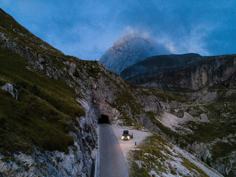 Aerial view of the Mangart saddle road leading into a tunnel, surrounded by rugged mountain peaks and rocky slopes under a twilight sky in Log pod Mangartom, Tolmin, Slovenia.