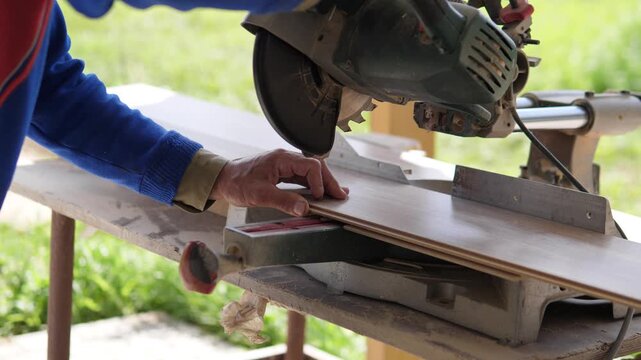 Close side view of worker hands guiding powered miter saw through wooden boards on rustic outdoor workbench, with blue sleeve, red vest edge, metal fence, blade housing, stacked