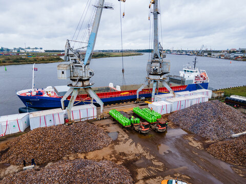 Riga, Latvia - 12 October 2023: Aerial view of a cargo ship being loaded with mineral fertilizers by large cranes at the port, with piles of bulk material and trucks nearby.