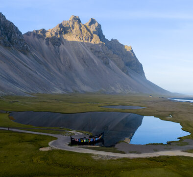 Aerial view of the dramatic Vestrahorn Mountain reflecting in a calm pond with a Viking ship replica moored on the shore in Hofn, Sveitarfelagid Hornaf, Iceland.