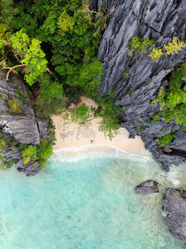 Aerial view of a couple lying on the sand in a starfish position on a beach at Shimizu Island surrounded by limestone cliffs and turquoise water in El Nido, MIMAROPA, Philippines.