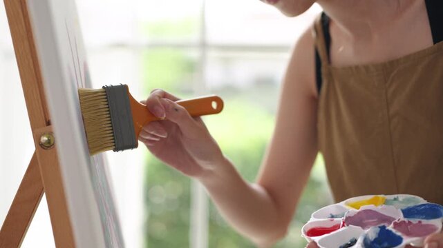 Creative young Asian woman artist painting on canvas in bright home studio. Relaxed female hobbyist enjoying peaceful indoor leisure time, expressing imagination and passion for art.