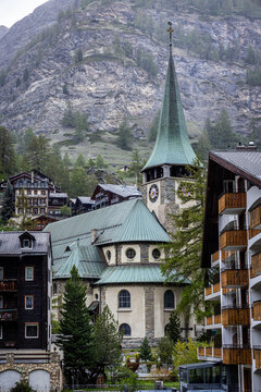 View of St. Mauritius Church with its distinctive green copper roof and clock tower nestled among traditional alpine buildings in Zermatt, Valais, Switzerland.