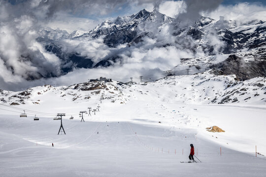 View of a skier in a red jacket on a snowy slope with the Matterhorn mountain in the background under a cloudy sky in Zermatt, Valais, Switzerland.
