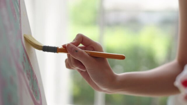 Creative young Asian woman artist painting on canvas in bright home studio. Relaxed female hobbyist enjoying peaceful indoor leisure time, expressing imagination and passion for art.