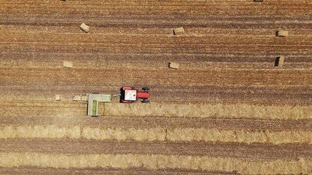 Aerial view of a red tractor pulling a baler through a harvested field with scattered hay bales in Jazak, Vojvodina, Serbia.