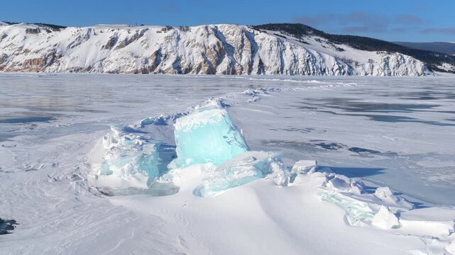 Aerial view of frozen Lake Baikal with large turquoise ice blocks and snow-covered mountains in the background in Khuzhir, Irkutsk Oblast, Russia.