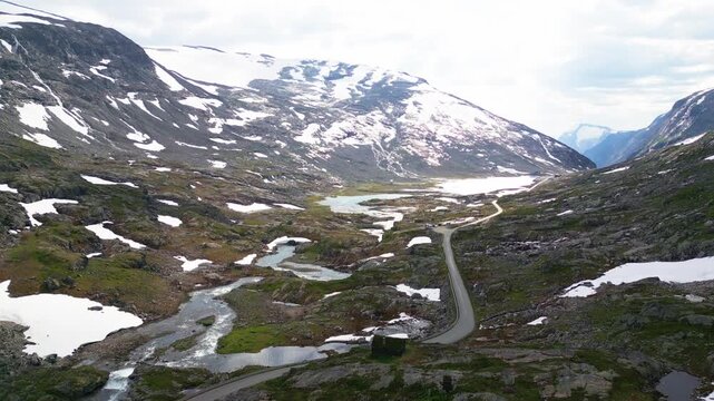 Aerial view of Old Strynefjell Mountain Road winding through a rugged mountain landscape with snow patches and a river in Stryn Municipality, Vestland, Norway.