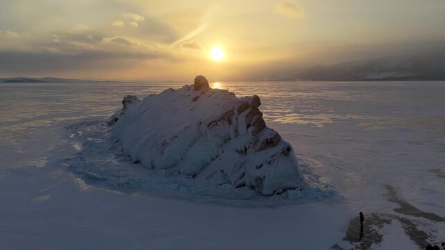 Aerial view of Shamanka Rock on frozen Lake Baikal at sunset with golden light reflecting on ice and people standing on the shore in Khuzhir, Irkutsk Oblast, Russia.