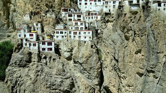 Aerial view of Phukthal Gompa monastery perched on a steep rocky cliff face with white buildings and green vegetation at the base in Phugtal, Himachal Pradesh, India.