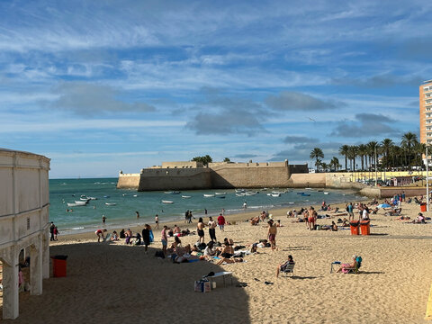 Cadiz, Spain - 25 October 2025: View of La Caleta beach with people relaxing on the sand, small boats in the water, and the historic stone fortifications of Castillo de Santa Catalina.