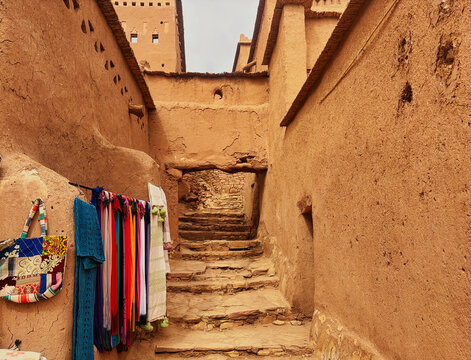 Ait Benhaddou, Morocco - 27 February 2026: View of Ait Benhaddou featuring traditional earthen architecture with stone steps between mud-brick walls and colorful textiles displayed.