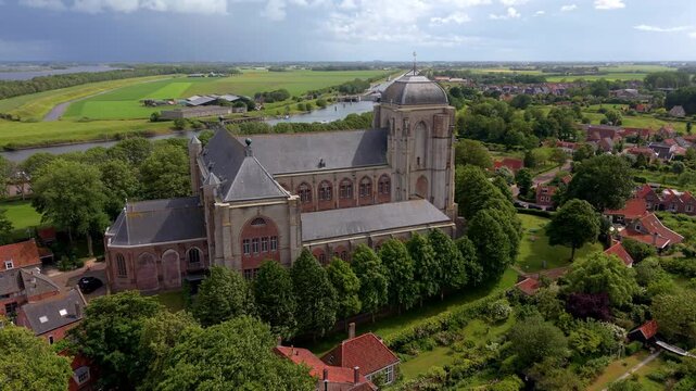 Aerial view video clip of historical Grote Kerk church, surrounding green trees, village houses, canal, and agricultural polders in Veere, Zeeland, Netherlands on a sunny day
