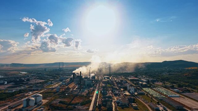 Huge disc of the sun dazzles the scenery of the modern plant. Drone footage over the industrial area producing smoke into the air.