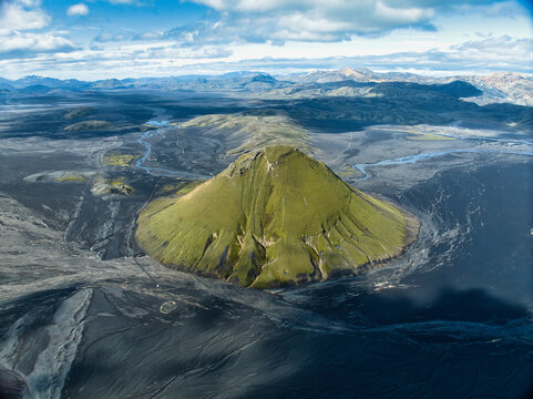 Aerial view of the vibrant green moss-covered Maelifell volcano rising from the vast black volcanic sand plains and glacial river patterns in Kirkjubaejarklaustur, Skaftarhreppur, Iceland.