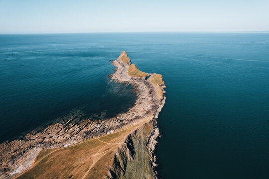 Aerial view of Worms Head Causeway extending into the sea with rugged rocky cliffs and grassy headlands in Rhossili, Wales, United Kingdom.