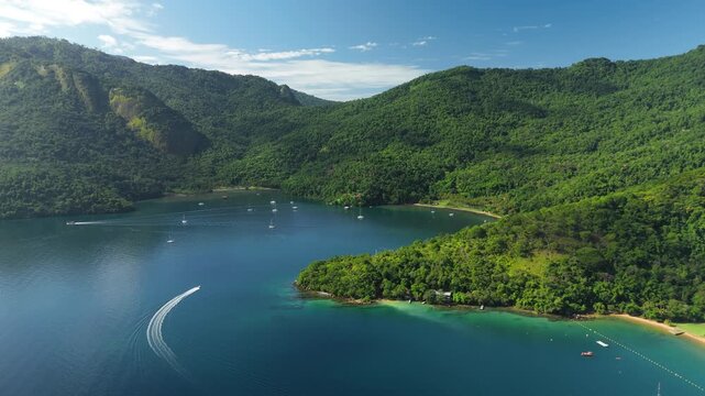 Motor boat moves fast on the calm and green lagoon with anchored sailing boats on the island of Ilha Grande, Brazil