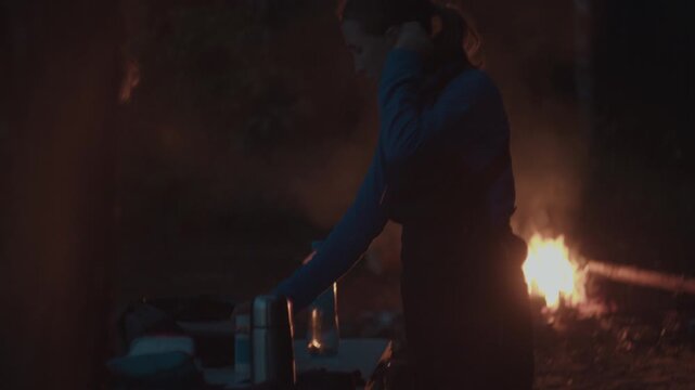 Woman hiker takes care of the camping site and does something with bonfire burning on the background