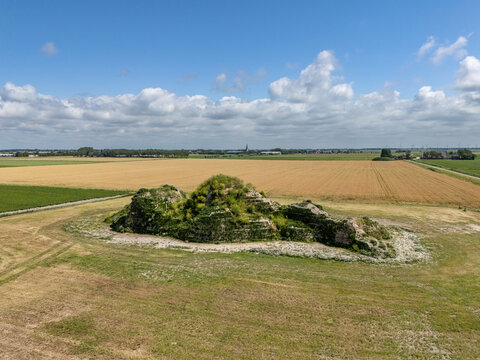 Aerial view of the Dijk van een wijf sculpture, a grassy mound structure set in a vast agricultural field under a bright blue sky with scattered clouds in Oosterbierum, Friesland, Netherlands.